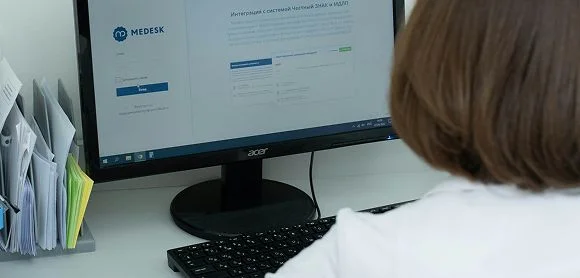 Eine Person mit kurzen braunen Haaren sitzt an einem Schreibtisch vor einem Computer mit geöffneter Praxismanagement-Software; links neben dem Monitor stehen Ordner mit Unterlagen.