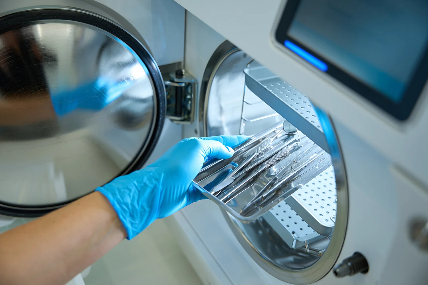 Eine behandschuhte Hand legt ein Tablett mit zahnmedizinischen Instrumenten aus Metall in ein Sterilisationsgerät mit offener Tür, um optimale Hygiene für die Patientenversorgung zu gewährleisten.