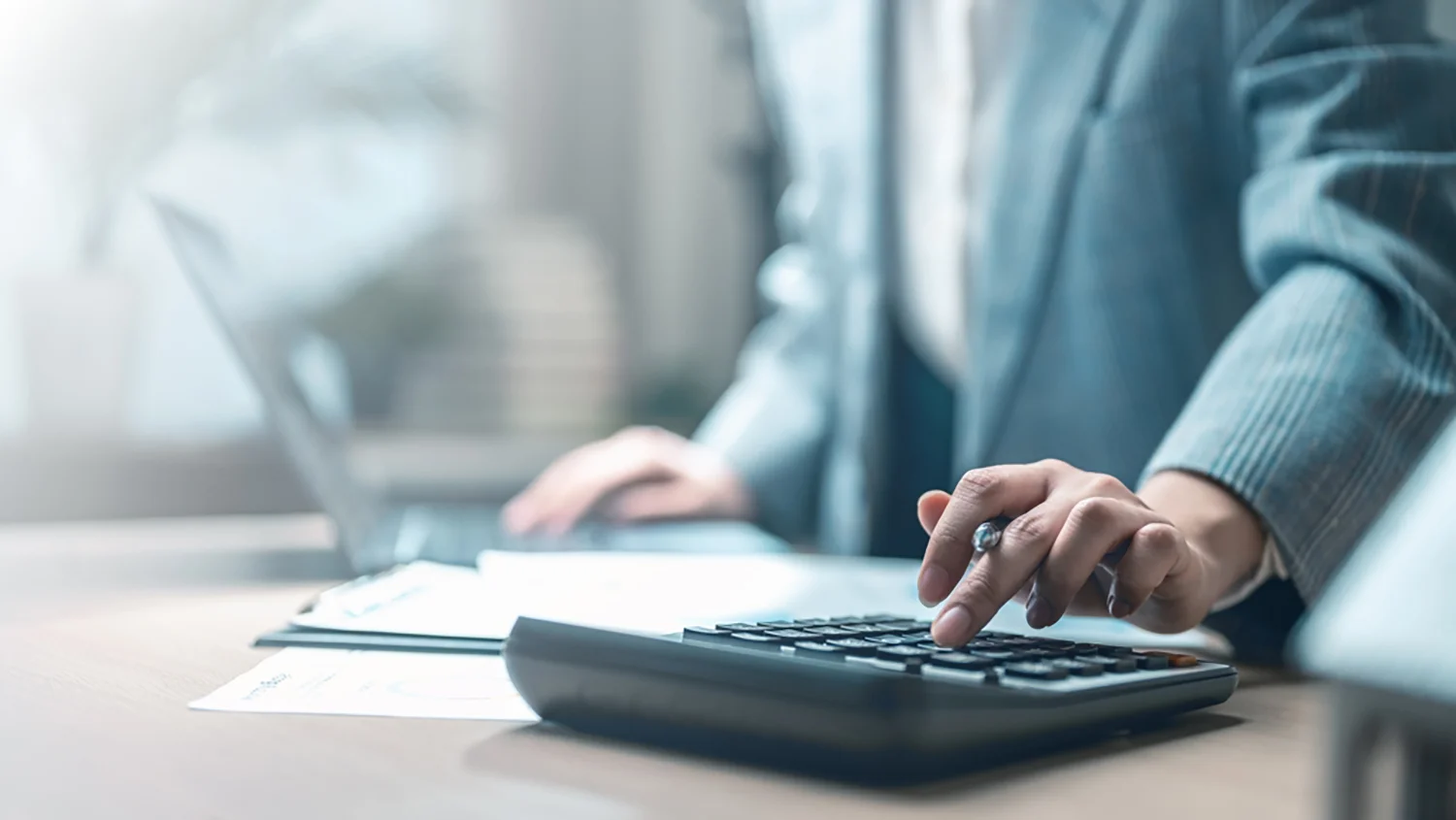 Eine Person im Anzug arbeitet an einem Schreibtisch mit Dokumenten, benutzt einen Taschenrechner und einen Laptop.