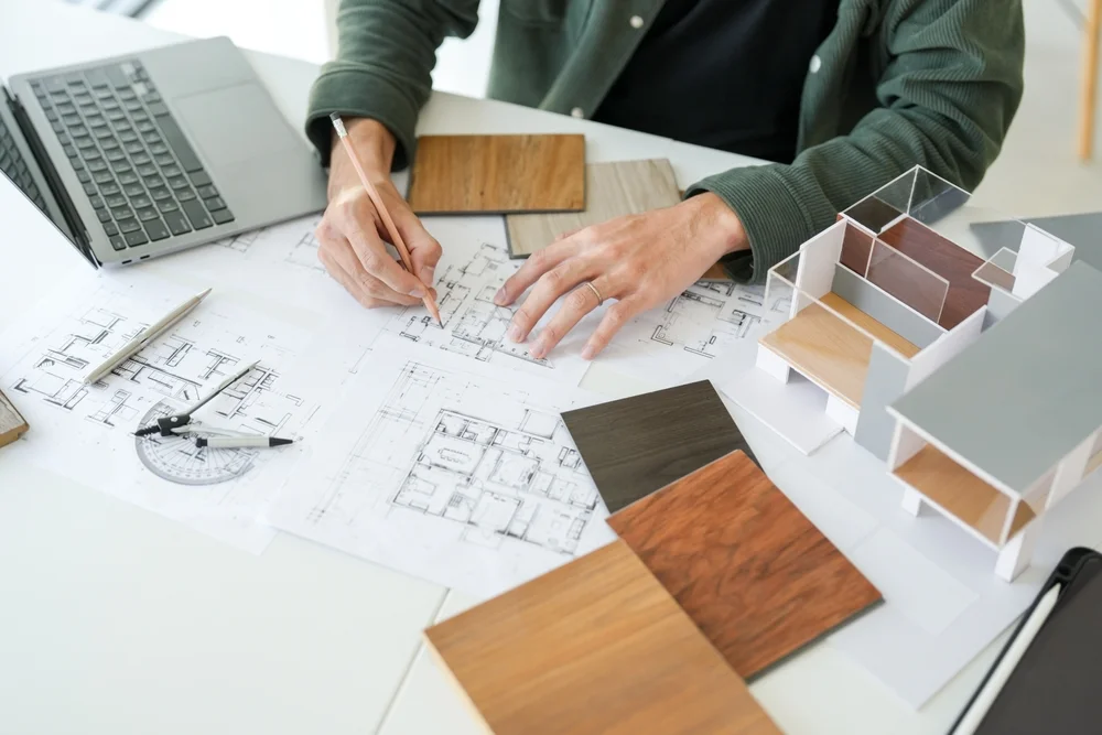 Ein Architekt arbeitet an einem Schreibtisch mit Bauplänen, Laptop, Holzmustern, einem maßstabsgetreuen Hausmodell und Zeichengeräten und macht sich Notizen zu den Grundrissen.