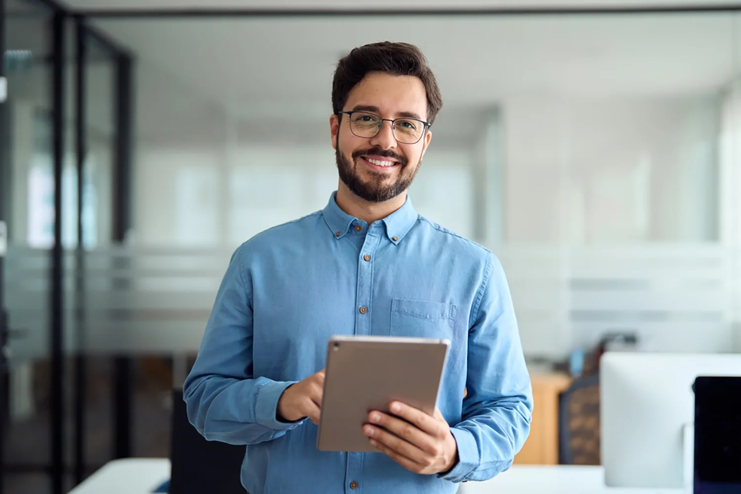 Ein lächelnder Mann mit Brille und Bart, der ein blaues Button-up-Hemd trägt, steht in einem modernen Büro, während er ein Tablet hält und benutzt.