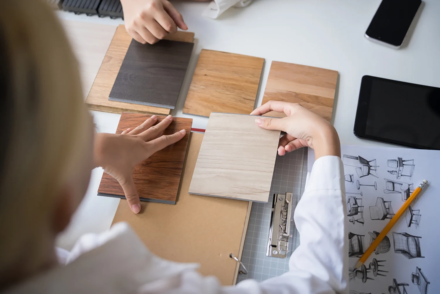 Zwei Personen begutachten Holzlaminate auf einem Tisch im Rahmen eines Praxisdesigns; daneben liegen Entwurfsskizzen, ein Bleistift, ein Klemmbrett und ein Tablet.