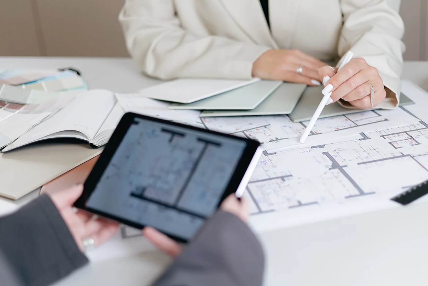 Zwei Personen prüfen architektonische Grundrisse an einem Tisch; eine hält ein Tablet mit Plänen, die andere zeigt mit einem Stift auf gedruckte Blaupausen.