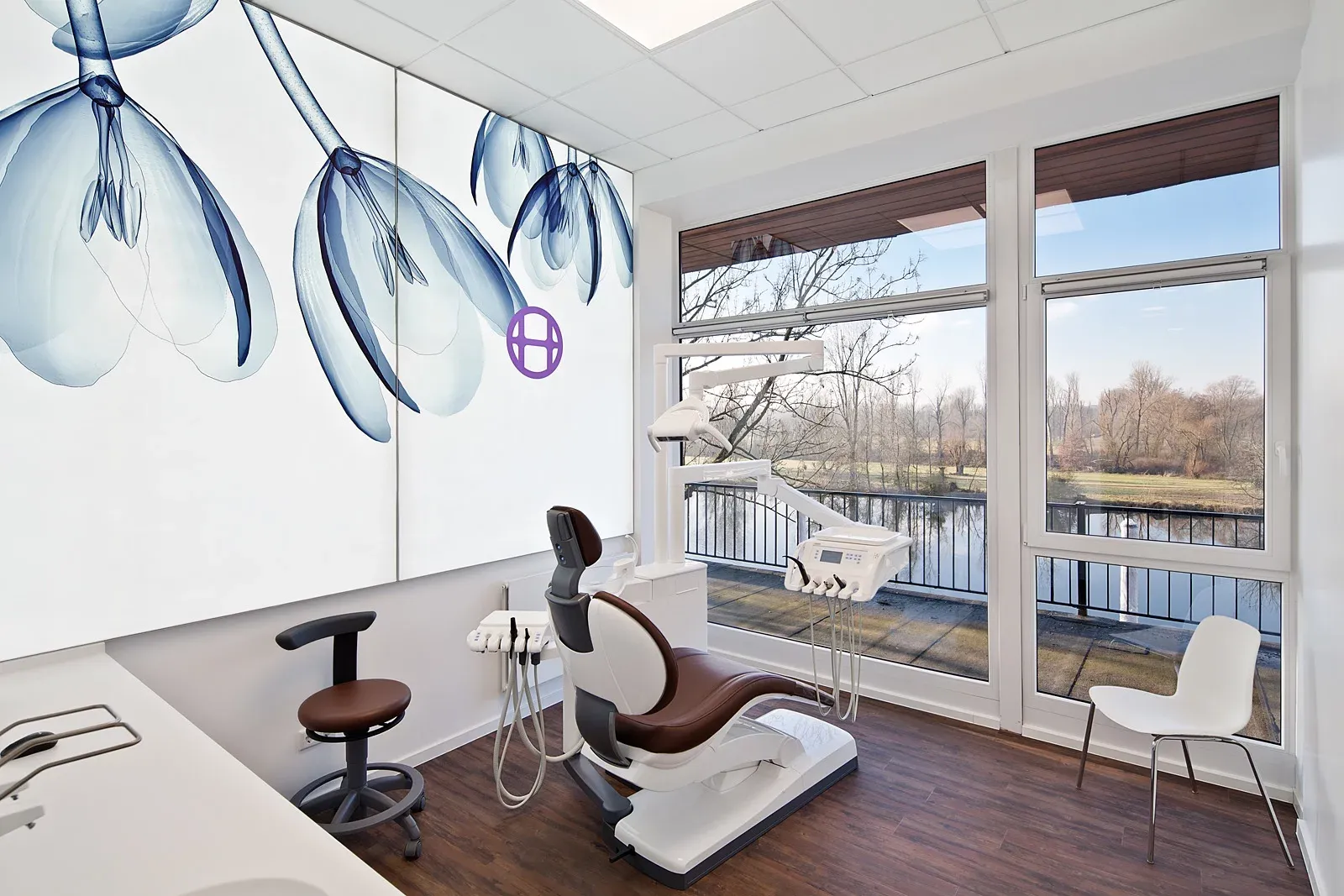 Behandlungszimmer mit Behandlungseinheit und Blick auf einen Fluss. An der Wand ein großes leuchtendes Wandbild mit floralem Design.
