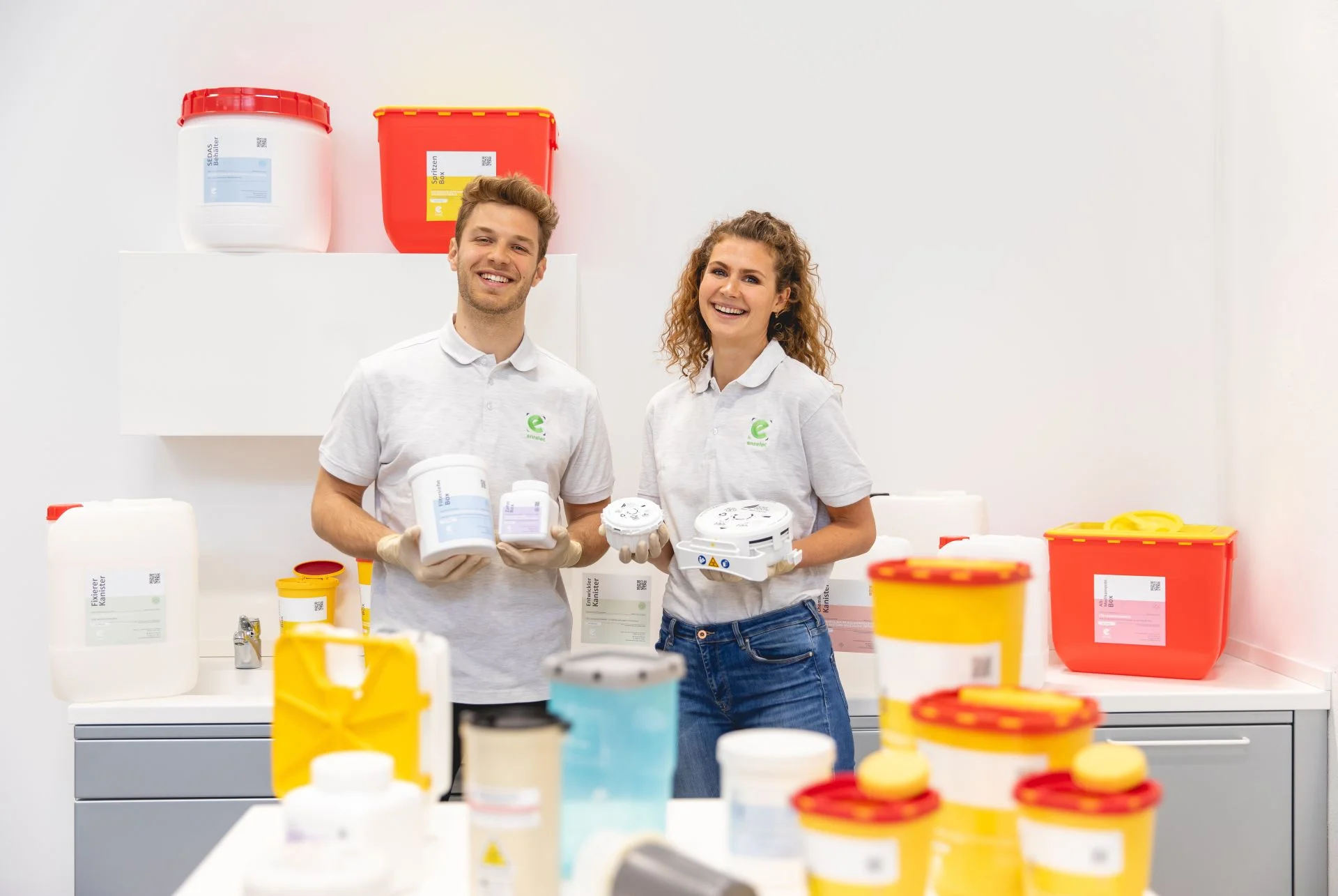 Zwei Mitarbeitende eines Entsorgungsdienstes stehen in einem Reinraum, lächeln und halten medizinische Abfallbehälter; im Hintergrund befinden sich farbige Behälter für medizinische Abfälle.