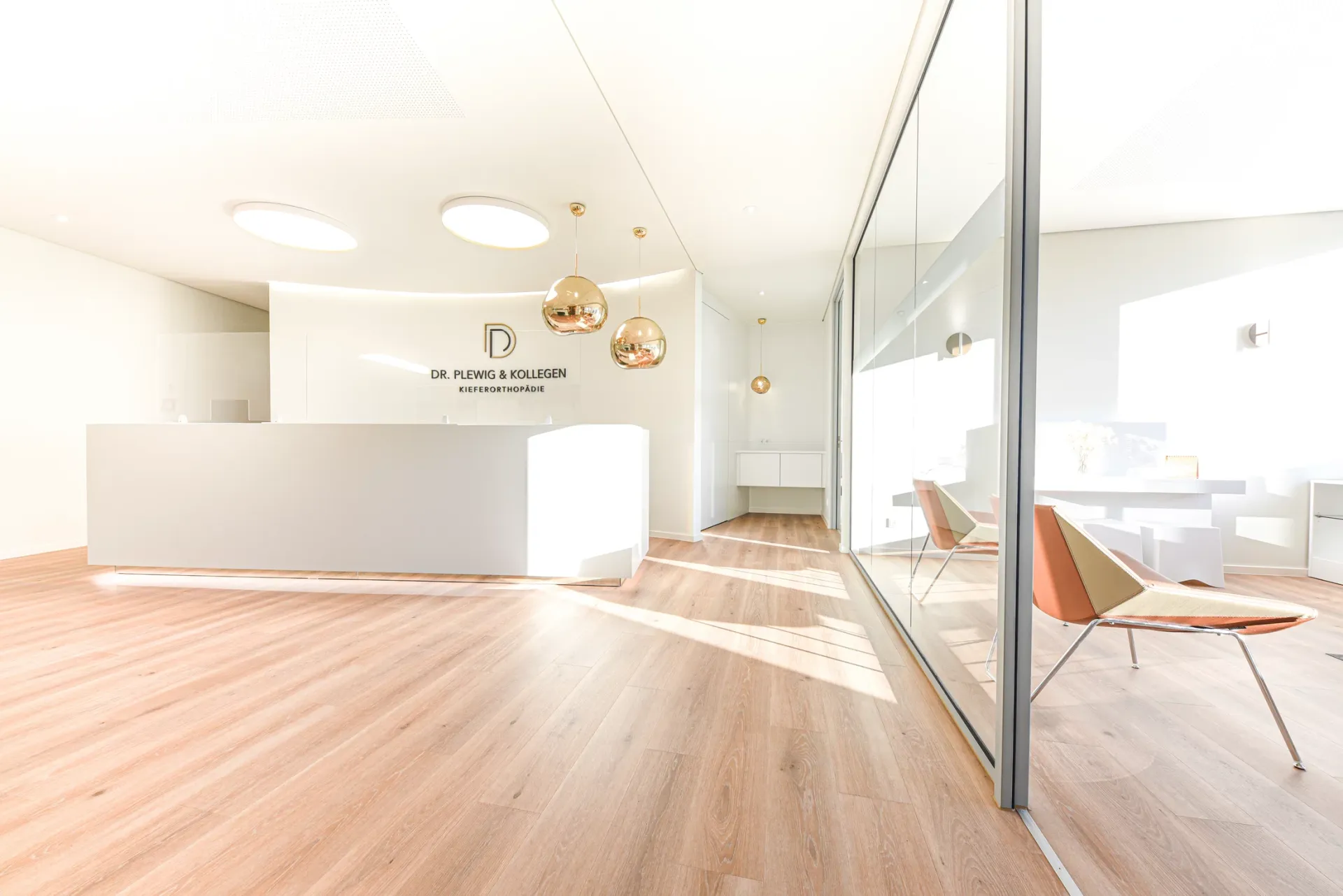 Empfangsbereich der KFO-Praxis mit moderner Ausstattung in weißen Farbtönen und hellbraunem Boden in Parkettoptik.