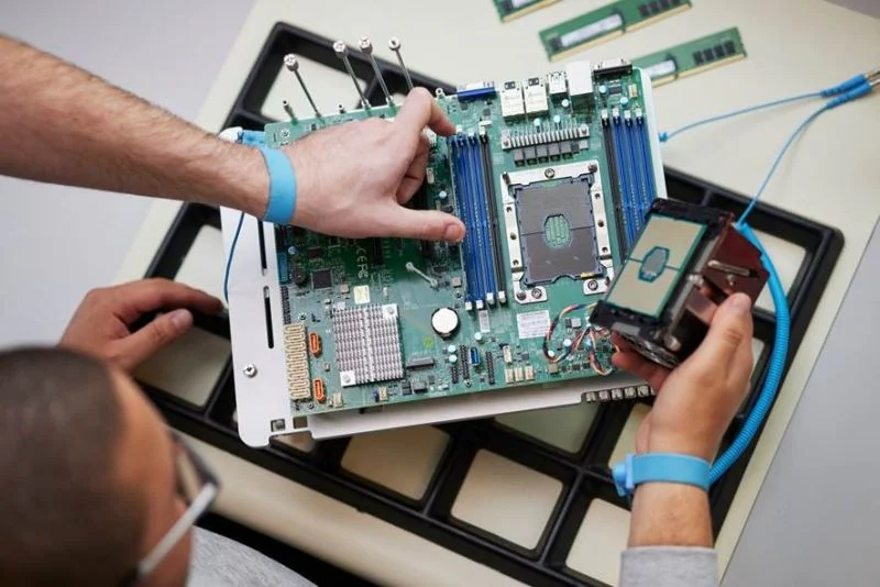 Zwei Personen montieren eine Computer-Hauptplatine mit antistatischen Handgelenksschlaufen; eine hält die Platine, die andere setzt eine CPU ein, während RAM-Sticks und Werkzeuge auf dem Tisch liegen.