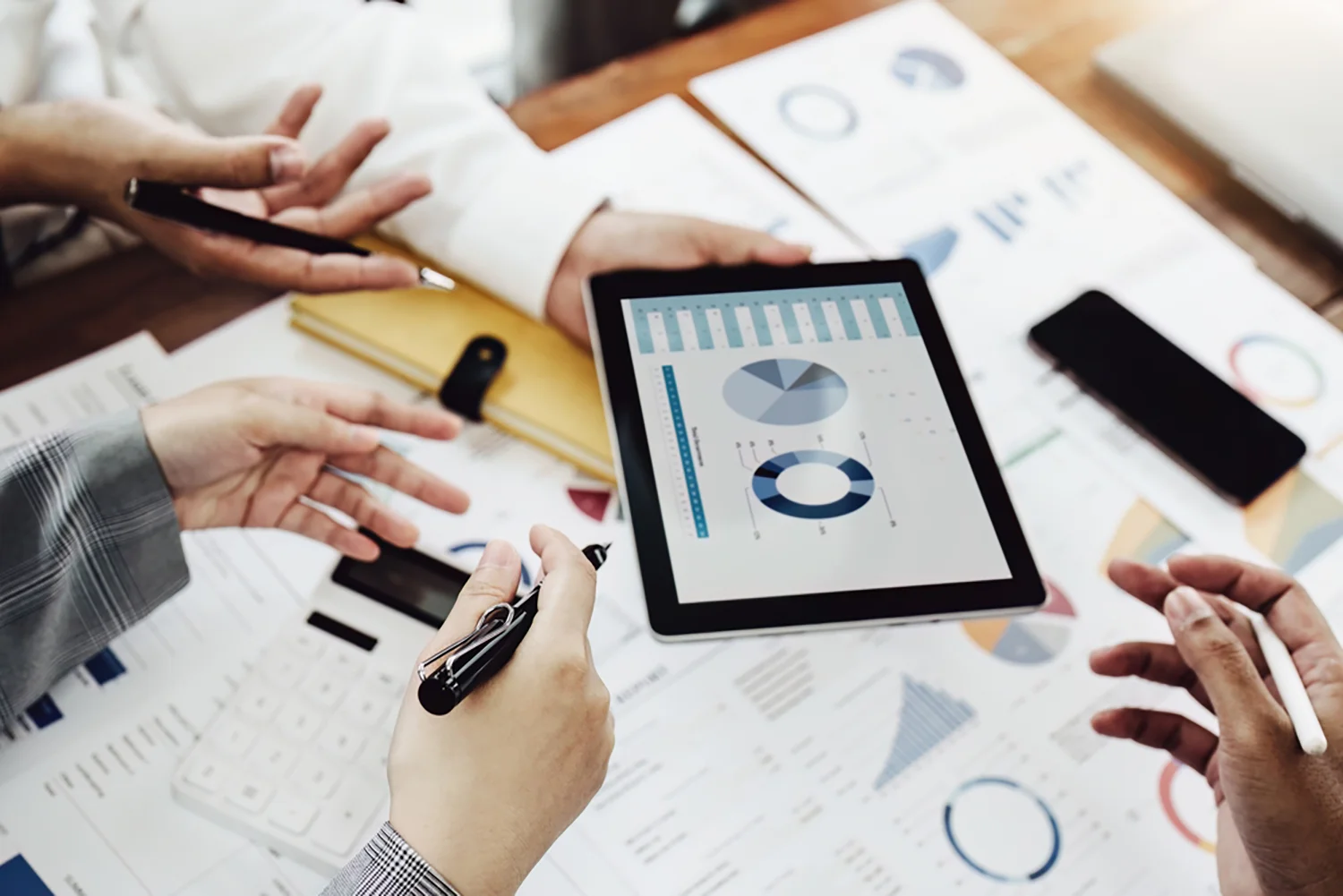 Vier Personen besprechen Daten auf einem Tablet mit Diagrammen und Grafiken; auf dem Tisch liegen Dokumente, ein Taschenrechner, ein Notizblock und ein Smartphone.