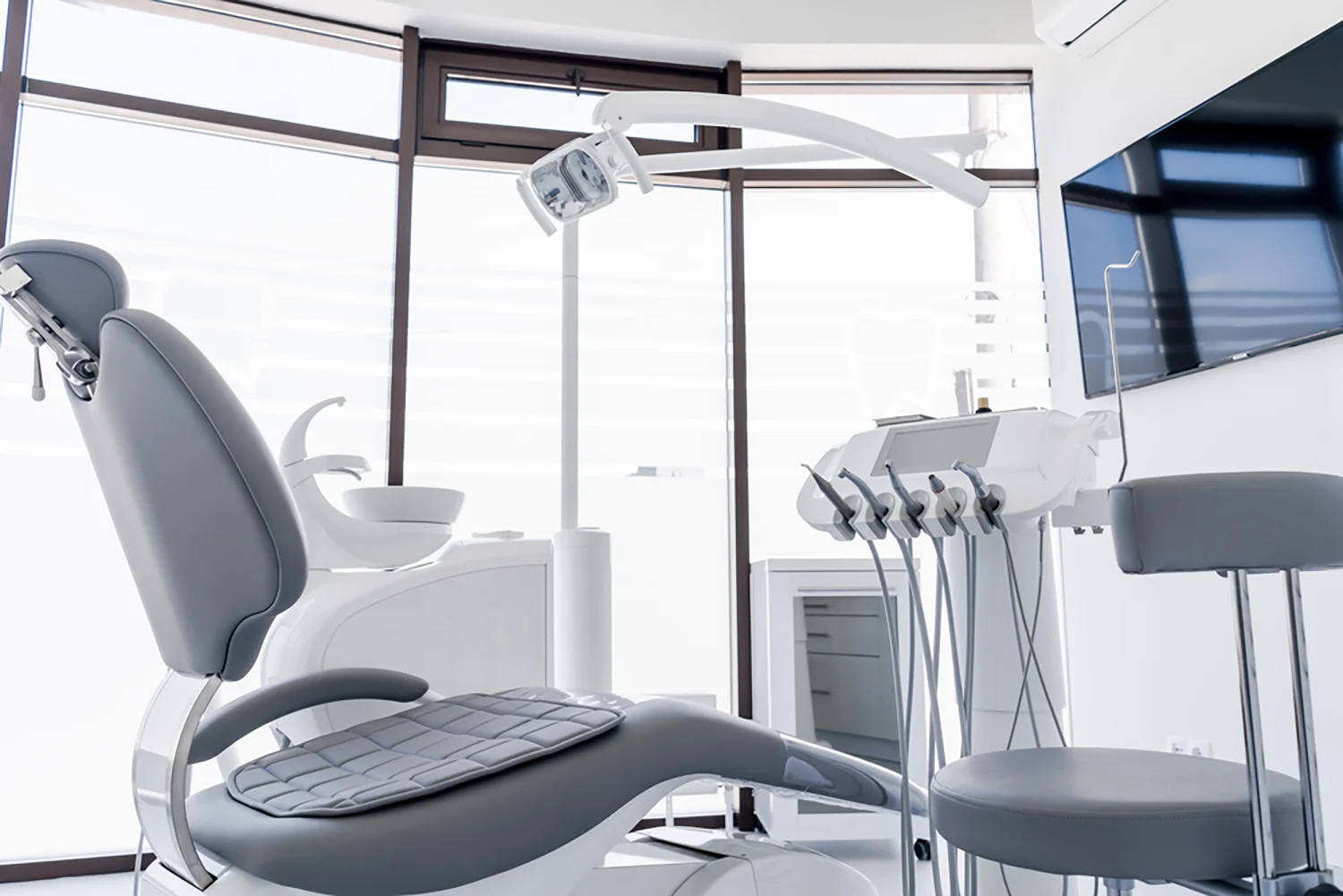 Moderne Zahnarztpraxis mit Behandlungsstuhl, Oberlicht, zahnärztlichen Instrumenten, Waschbecken und Flachbildfernseher; große Fenster sorgen für Tageslicht.