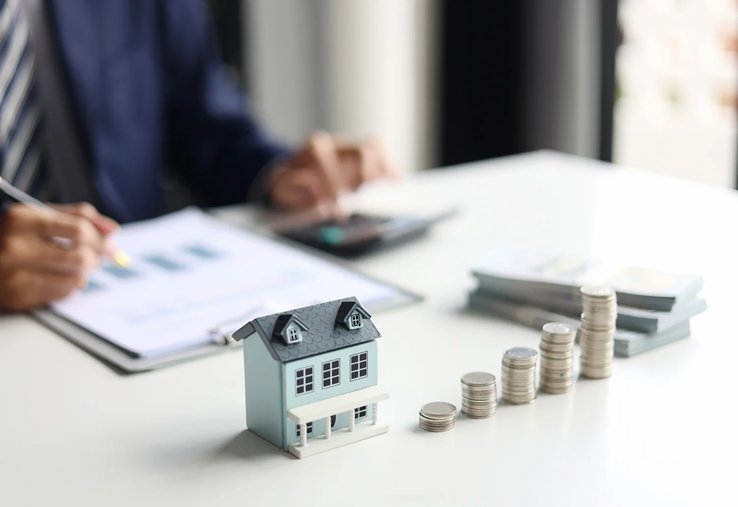 Ein kleines Modellhaus und ein Stapel Münzen stehen auf einem Schreibtisch, während eine Person im Anzug einen Taschenrechner benutzt und Dokumente überprüft.