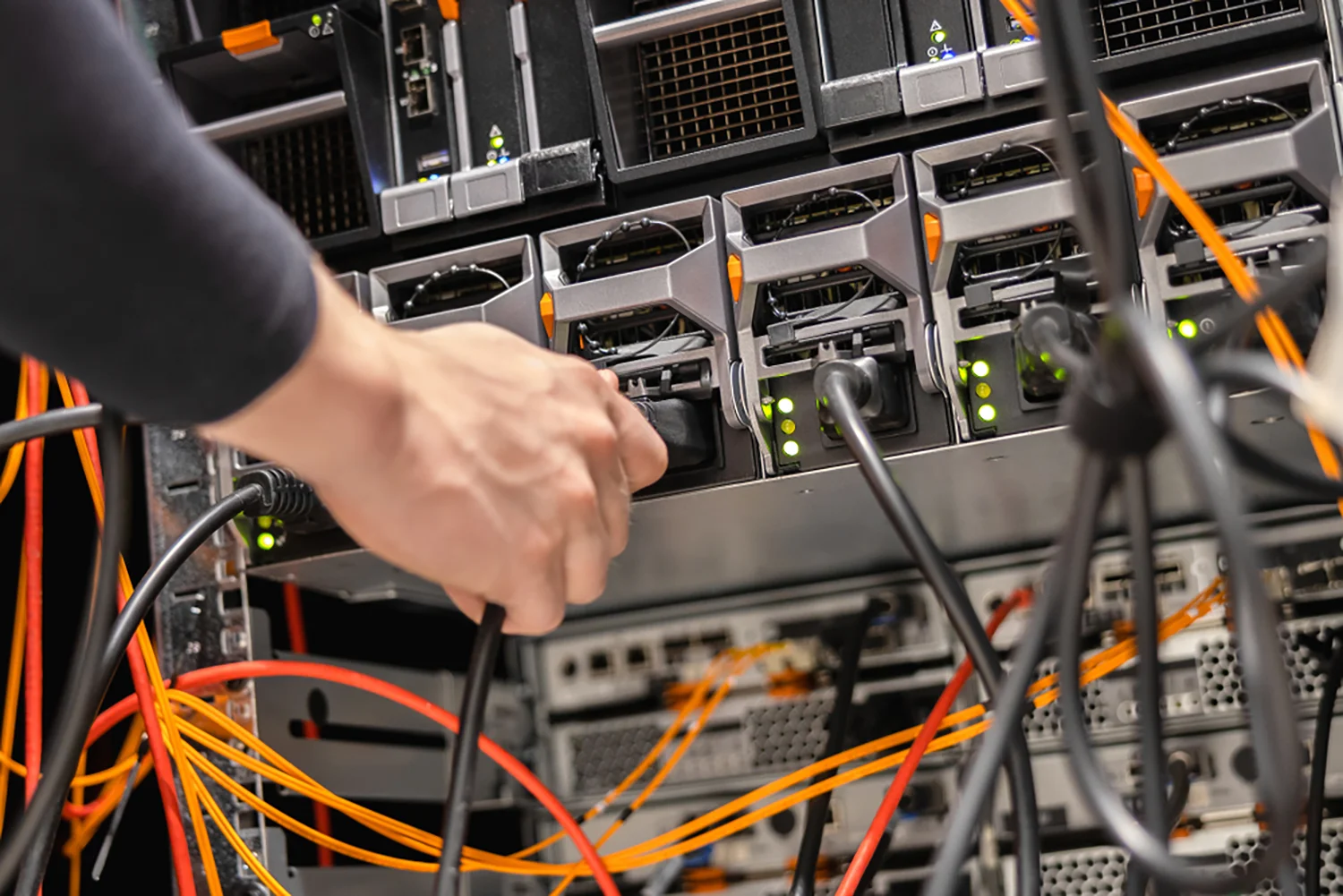 Eine Hand verbindet ein Kabel an einem Netzwerkserver-Rack mit eingesteckten orangefarbenen und schwarzen Kabeln sowie sichtbaren Kontrollleuchten.